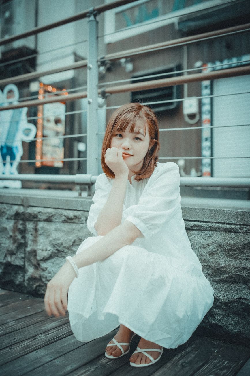 A serene adult woman in a white dress sits outdoors, smiling gently in an urban setting.