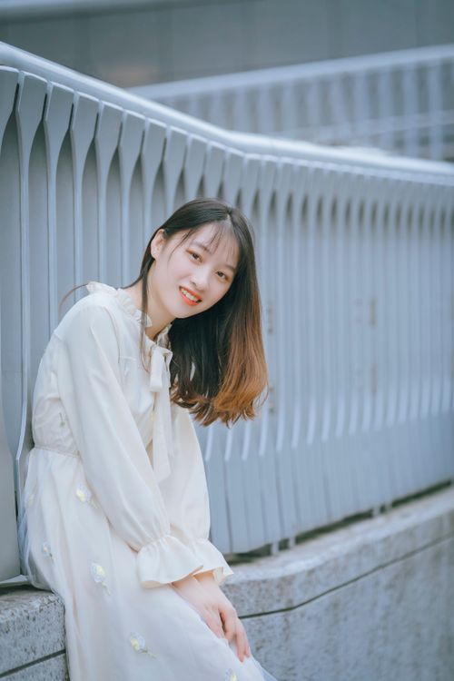 Free Asian woman in a white dress sits smiling by metal railings outdoors. Stock Photo