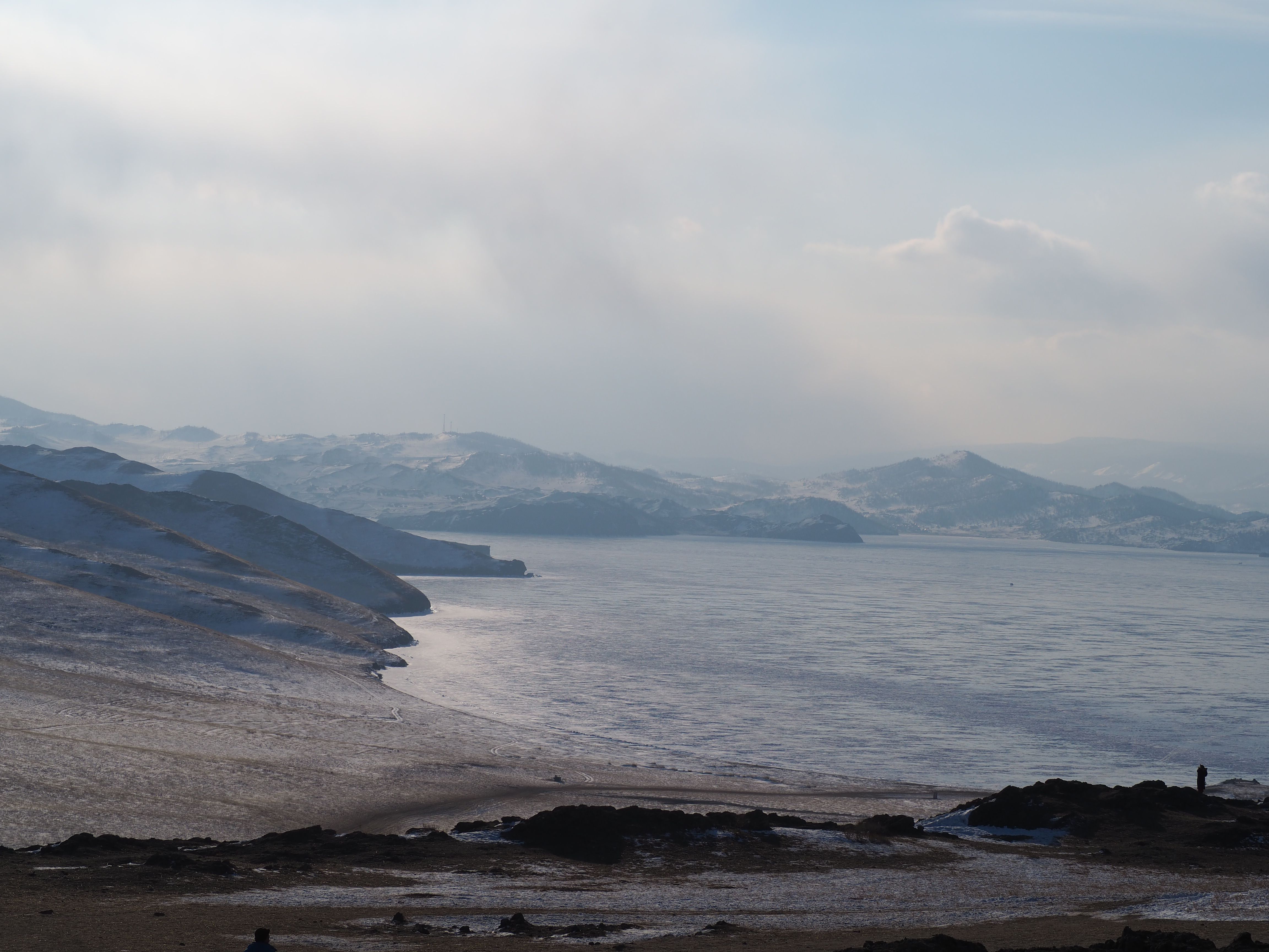 Calm winter scene featuring a snow-covered shoreline with distant mountains under a cloudy sky.