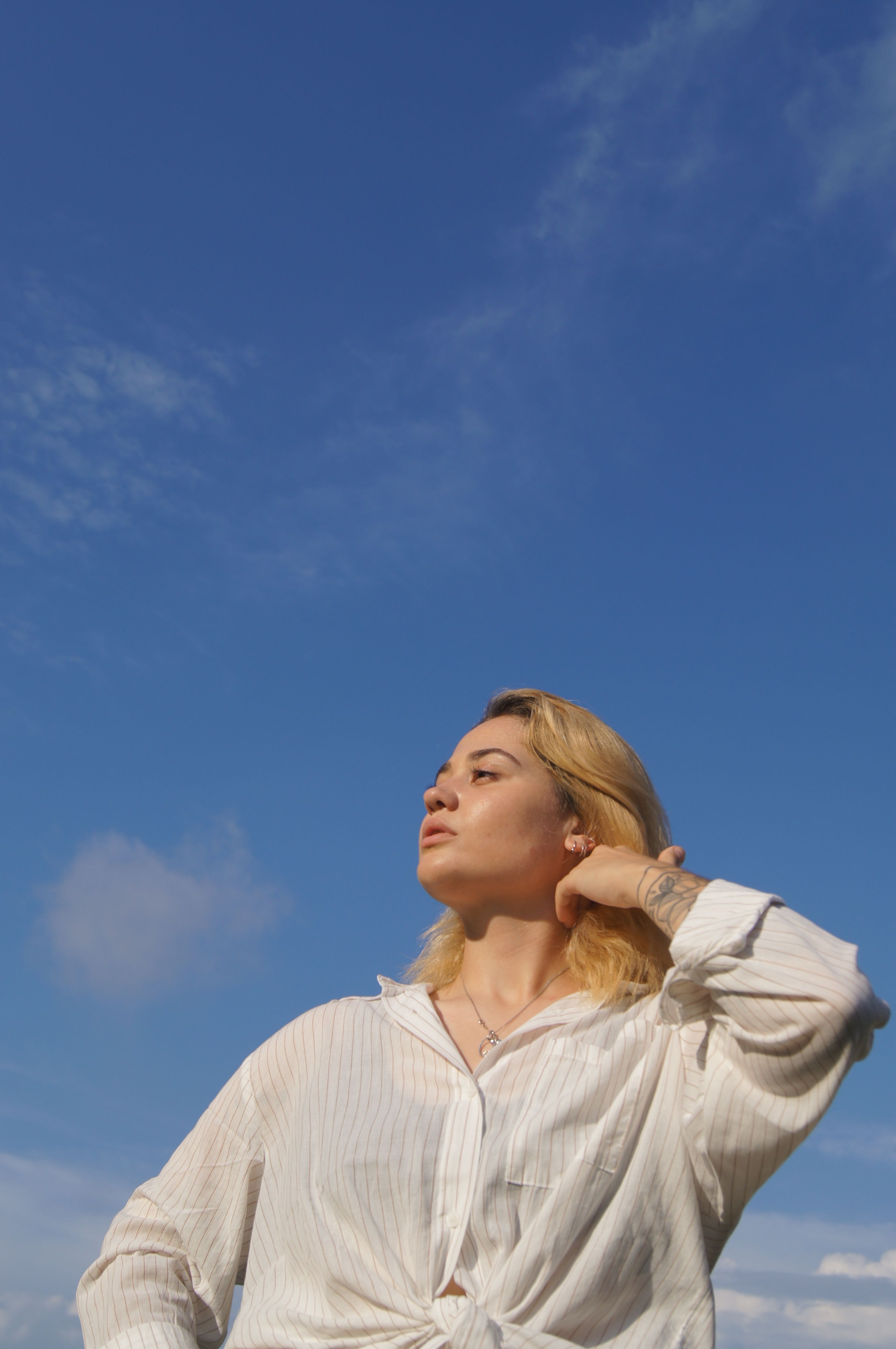 A confident woman poses outdoors in a white shirt against a clear blue sky. Low angle shot.