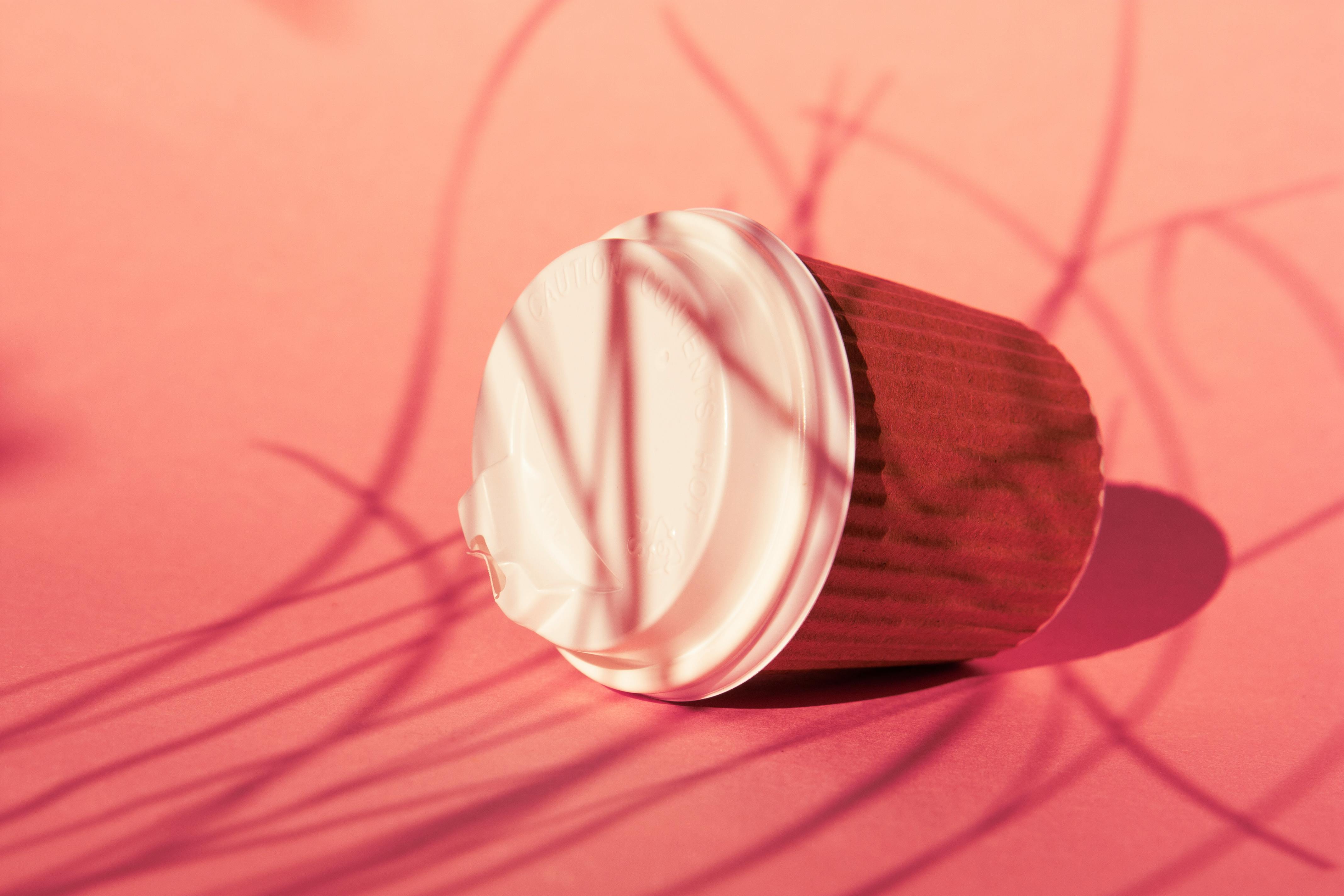 Stylish disposable coffee cup with lid, casting shadows on a pink background.