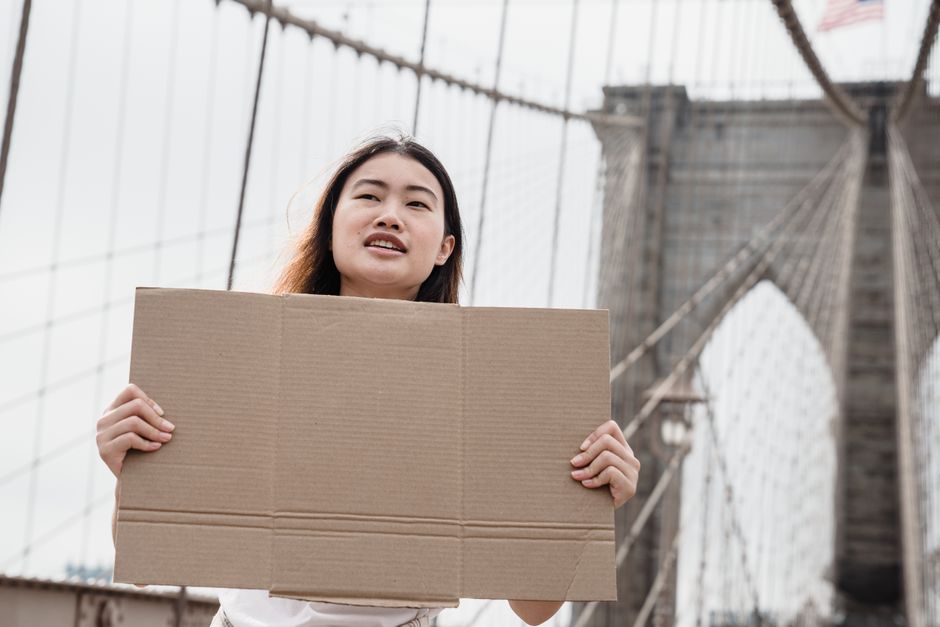 Asian woman holds a blank cardboard sign on Brooklyn Bridge, symbolizing protest and activism.