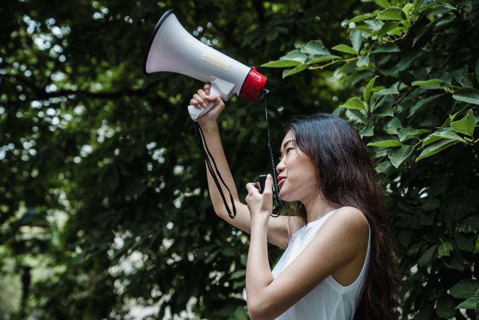A young woman passionately speaks into a megaphone in a lush green outdoor setting.
