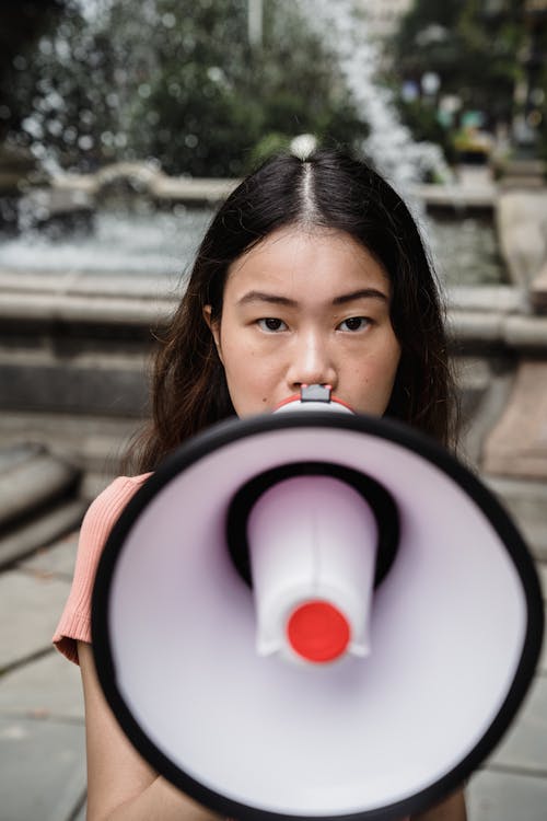 Free Close-up of an Asian woman holding a megaphone in an outdoor setting with a water fountain. Stock Photo