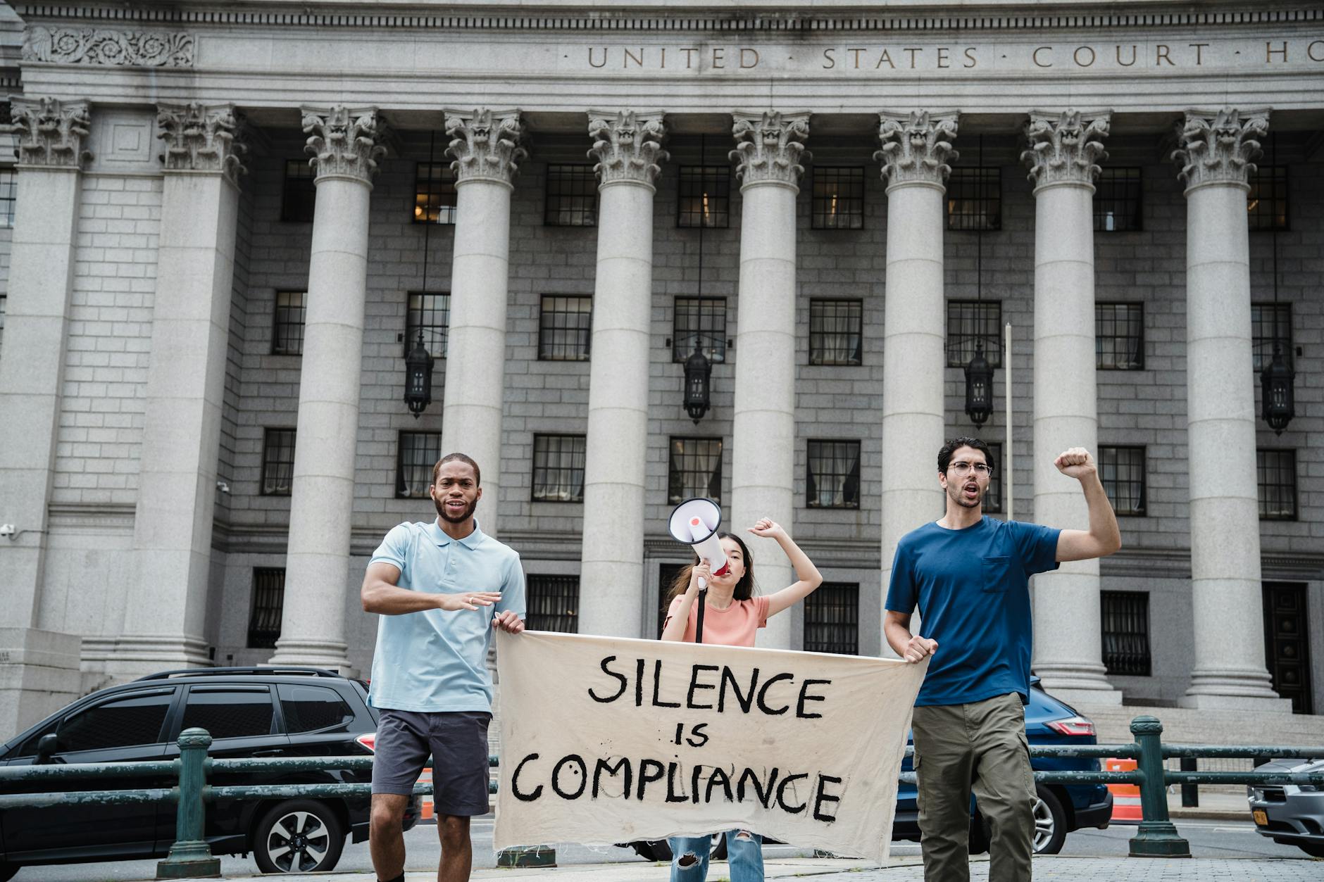 Three protesters with a 'Silence is Compliance' banner in front of a courthouse.