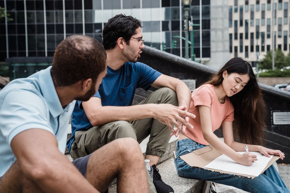 Tres jóvenes discutiendo ideas mientras están sentados en una escalera al aire libre. Ambiente colaborativo y creativo.