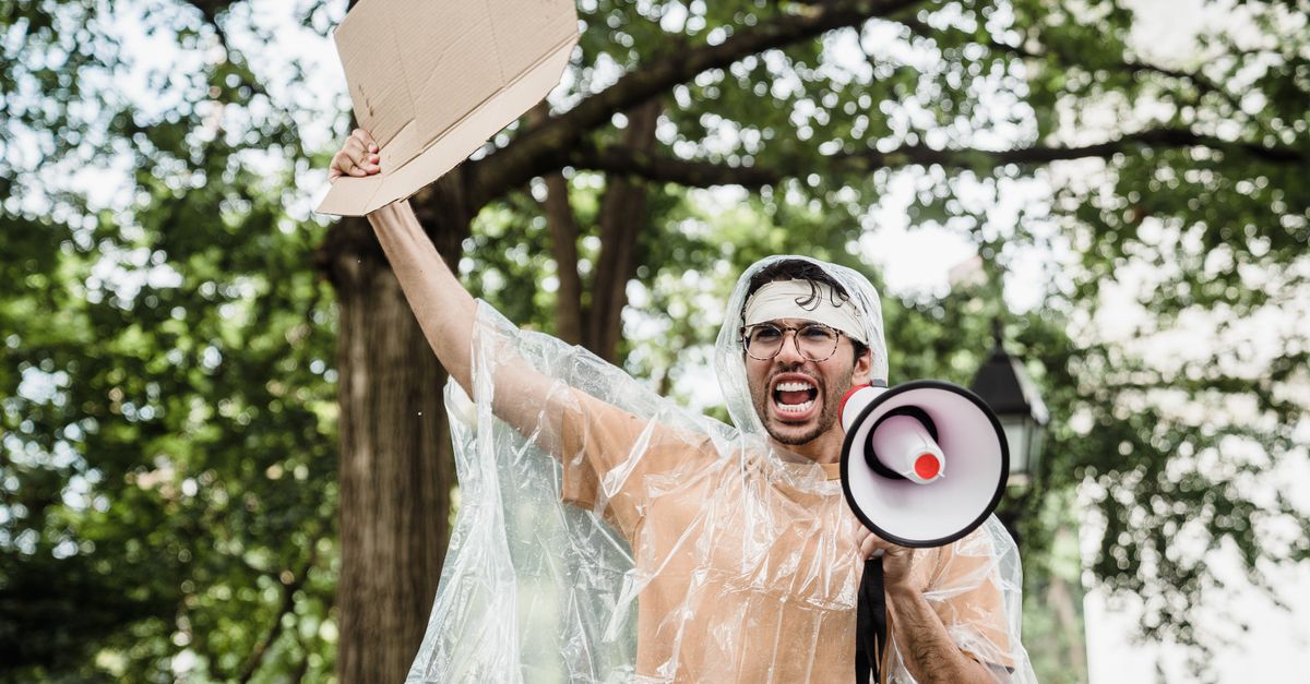 Man holding megaphone and cardboard at protest rally in park.