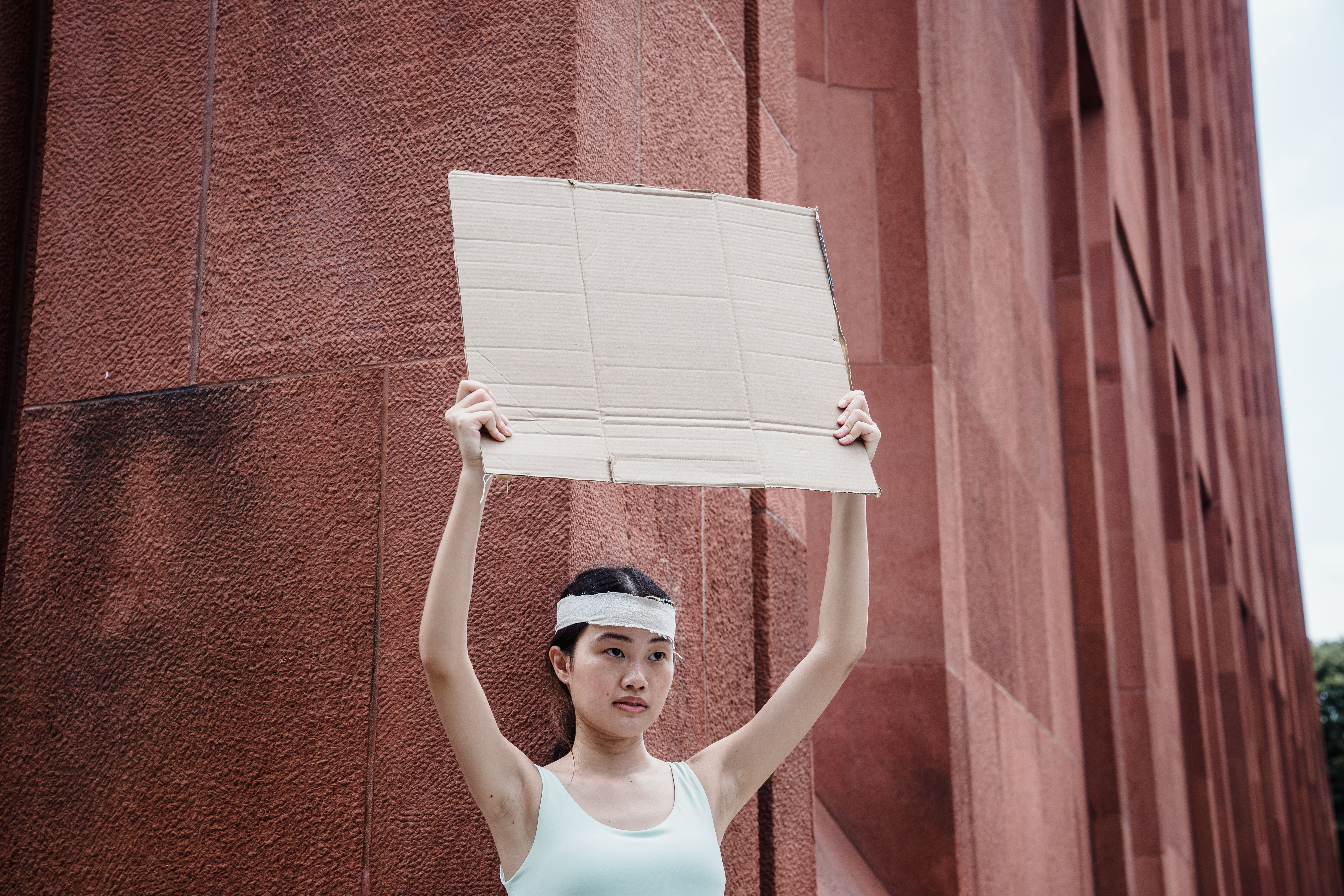 A young woman holds a blank cardboard sign in an urban setting, suggesting protest or demonstration.