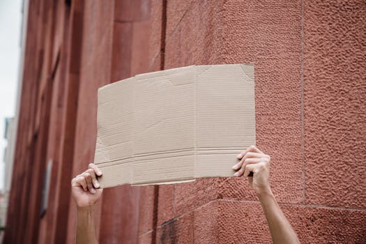 A person holds a blank cardboard sign in front of a red brick wall, symbolizing activism and protest.