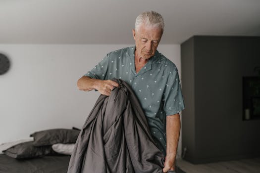 Elderly man with gray hair adjusting a bed. Indoor lighting sets a calm tone.