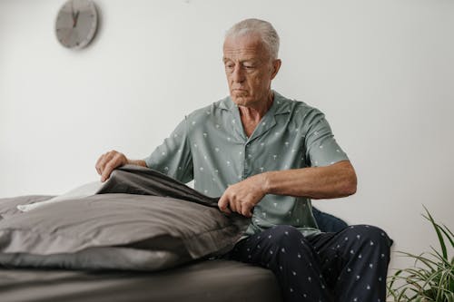 An Elderly Man in Gray Shirt and Black Pants Sitting on the Bed