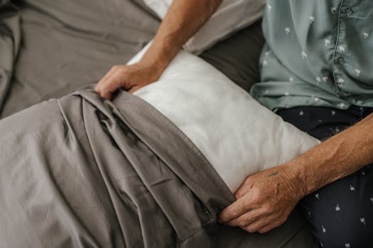 Close-up of a senior adult carefully arranging a bed with grey sheets and a white pillow.