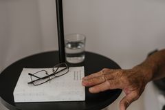 Close-up of an elderly hand reaching for glasses on a bedside table with a book and water glass, in a calm setting.