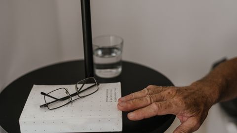 Close-up of an elderly hand reaching for glasses on a bedside table with a book and water glass, in a calm setting.