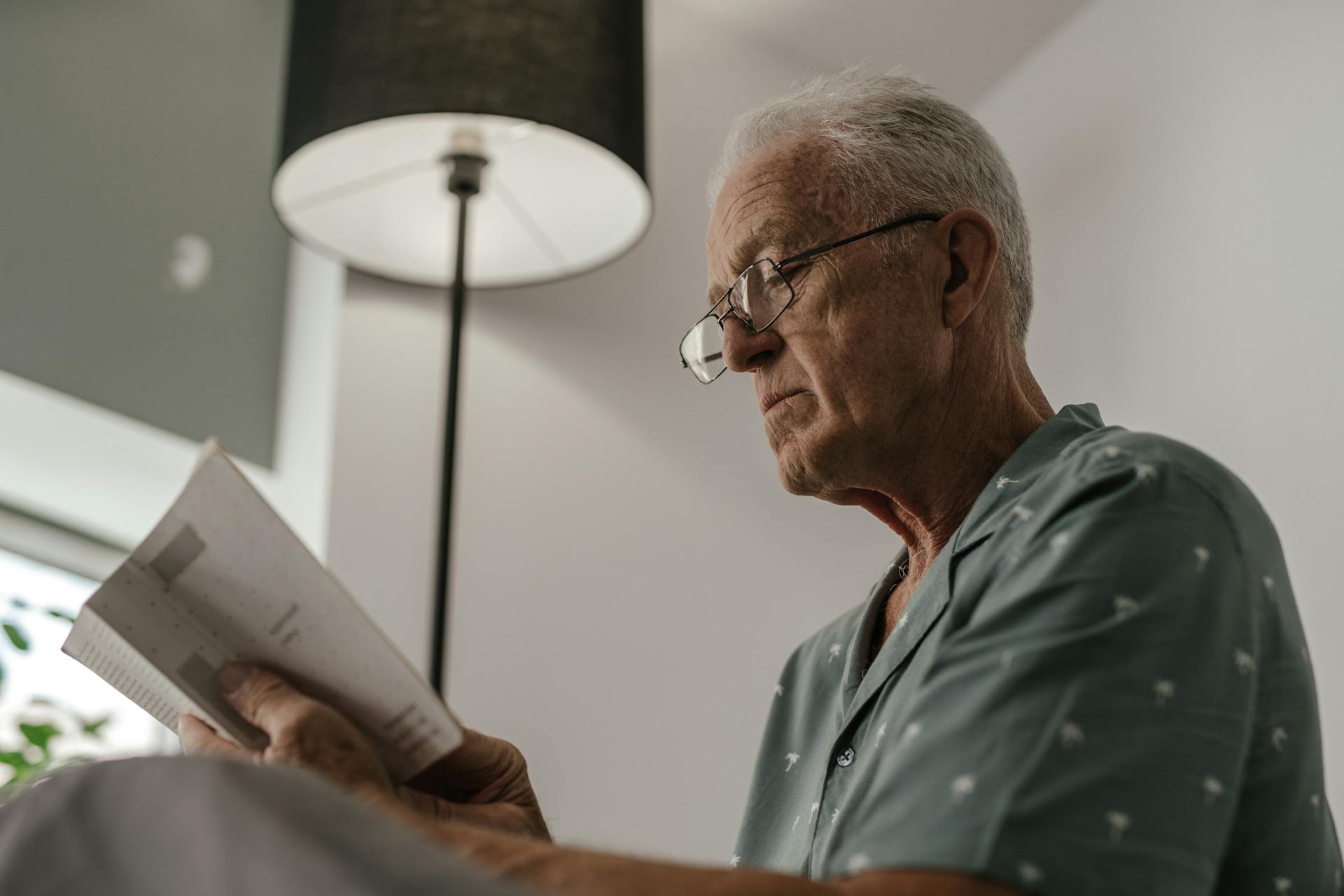 Senior man with eyeglasses reading a book under a lamp in a comfortable and serene indoor setting.