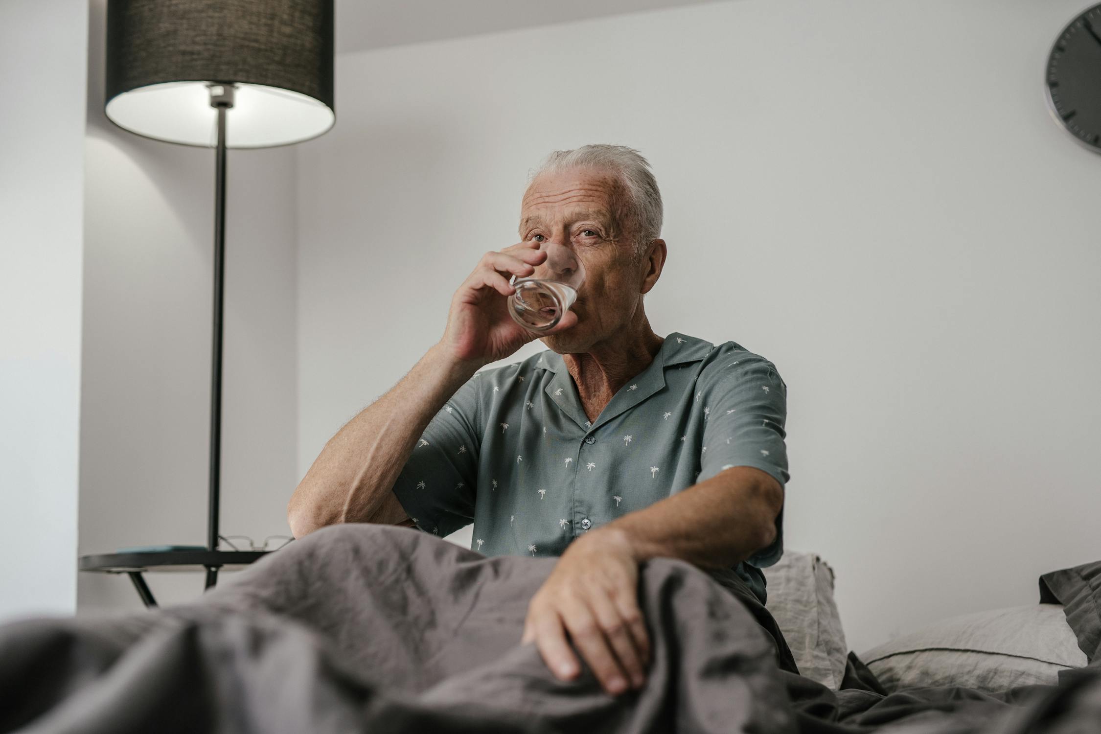 An Elderly Man Drinking a Glass of Water while Sitting on the Bed