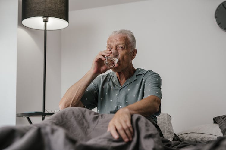 An Elderly Man Drinking A Glass Of Water While Sitting On The Bed