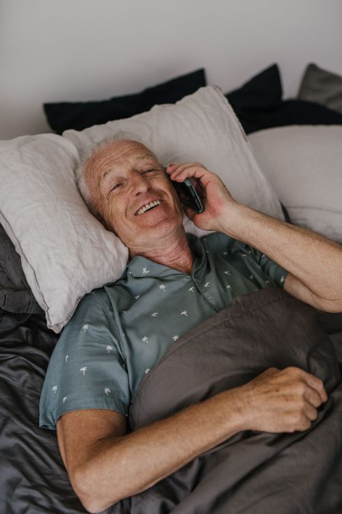 Free Senior man lying in bed, smiling and talking on a smartphone, exuding a sense of joy and comfort. Stock Photo