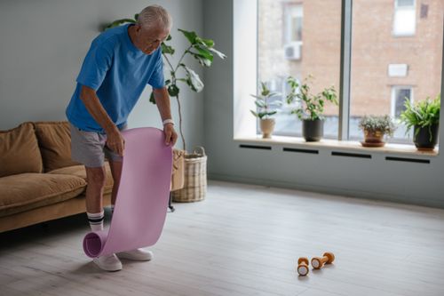Free A senior man unfolds a yoga mat indoors, preparing for a fitness routine with dumbbells nearby. Stock Photo