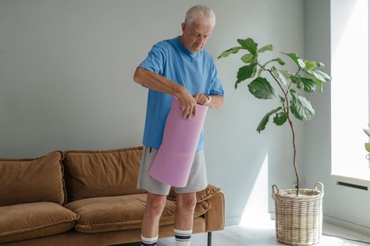 Elderly man in cozy room rolling out yoga mat for a healthy lifestyle workout.