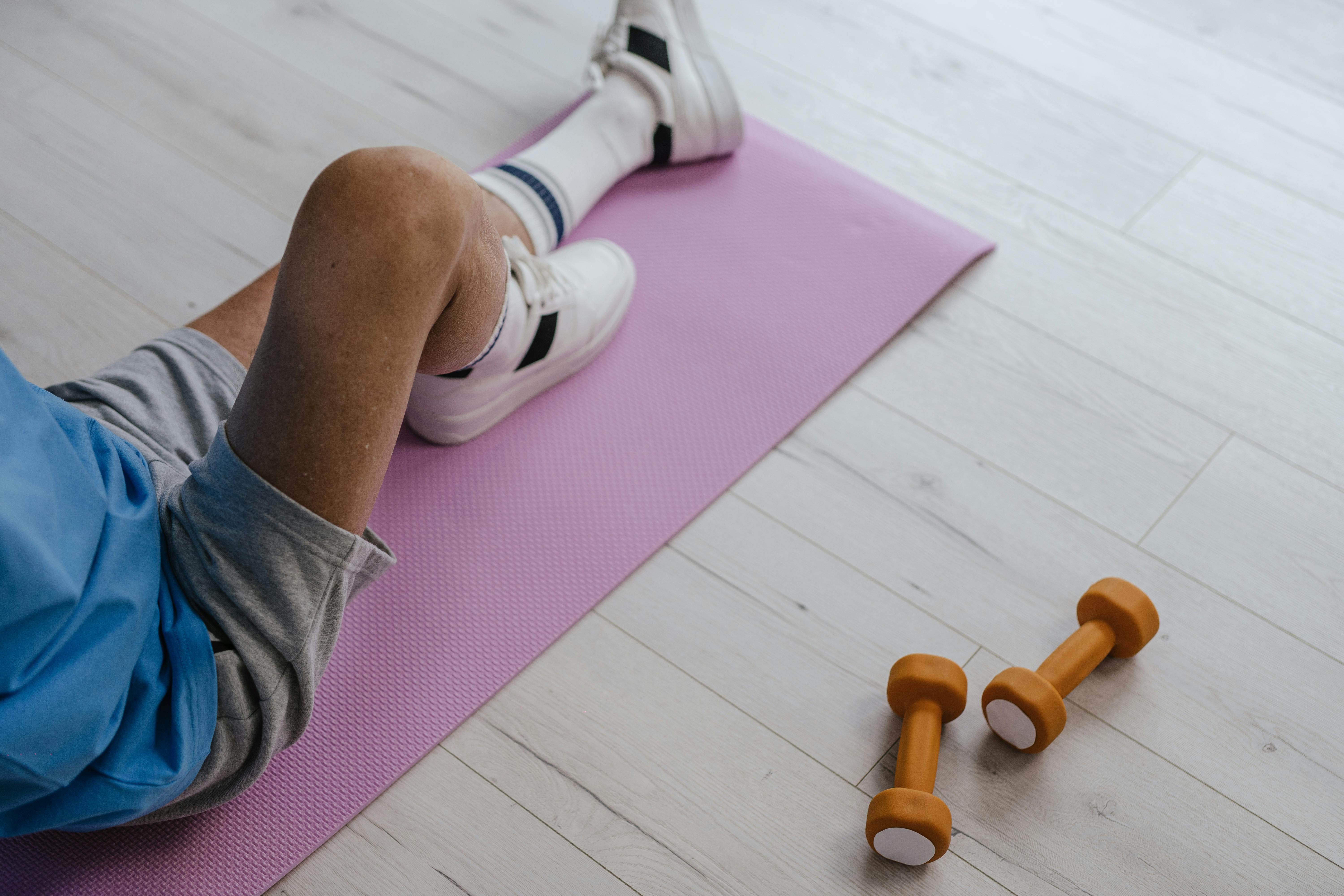 Senior adult sitting on a pink yoga mat indoors with orange dumbbells, promoting a healthy lifestyle.