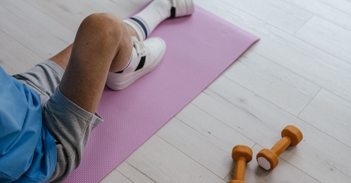 Senior adult sitting on a pink yoga mat indoors with orange dumbbells, promoting a healthy lifestyle.