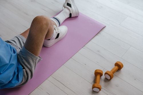 Free Senior adult sitting on a pink yoga mat indoors with orange dumbbells, promoting a healthy lifestyle. Stock Photo