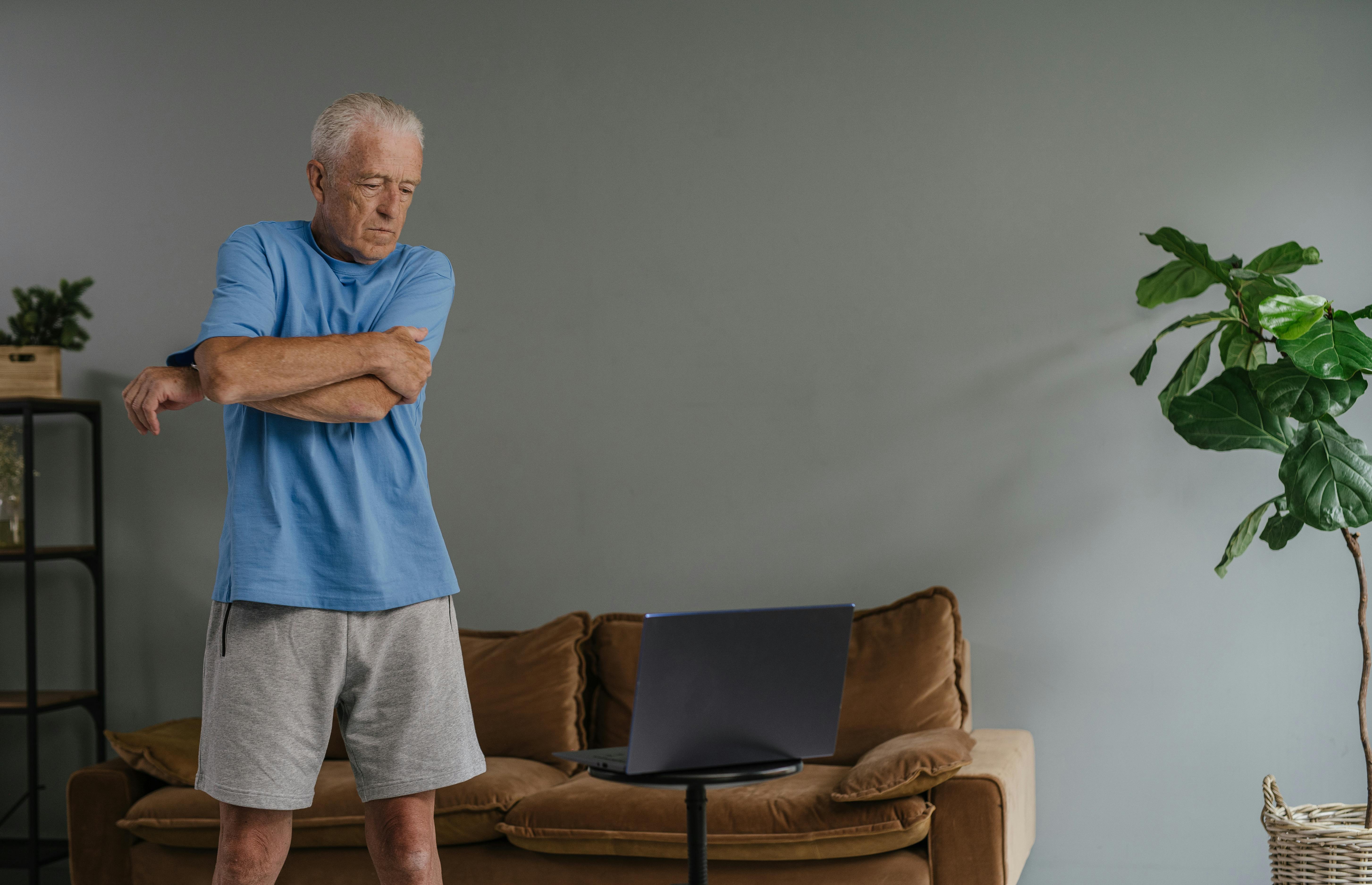 Senior man in casual wear doing a gentle stretch near a laptop in a cozy living room setting.
