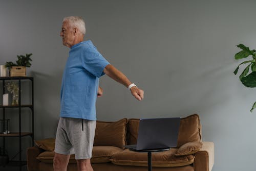 Man in Blue Crew Neck T-shirt and Gray Shorts Stretching Beside Brown Sofa