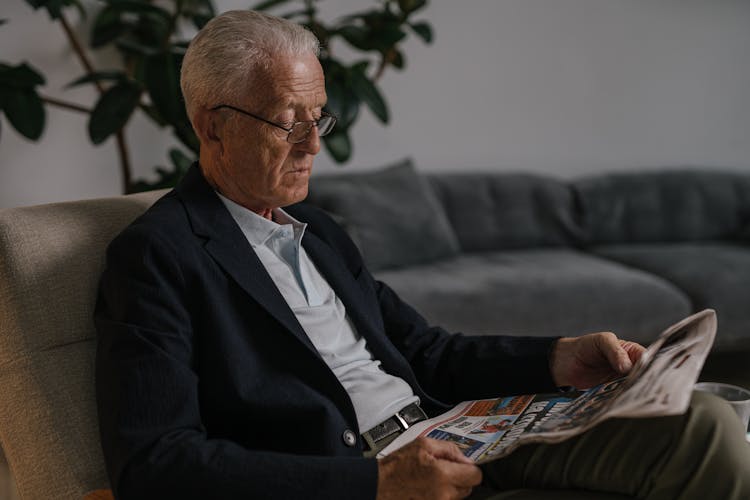 An Elderly Man In Black Suit Sitting On The Couch While Reading Newspaper