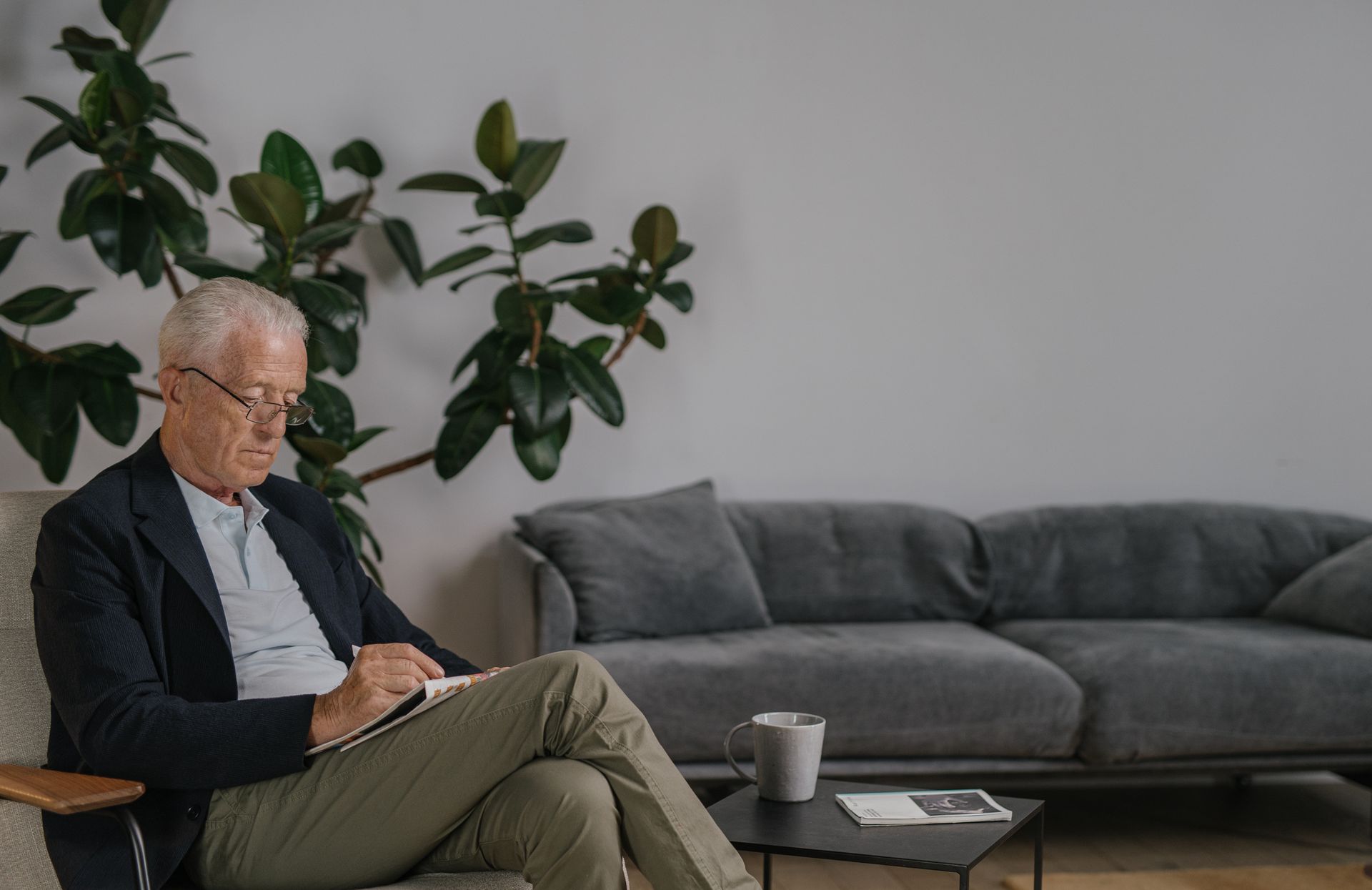 Elderly man writing in a modern, minimalist living room with a plant and sofa.