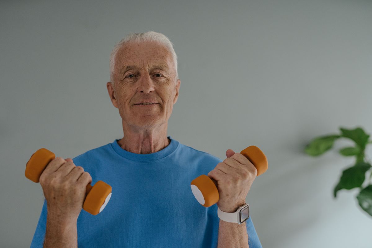 A man in his 60s at home, smiling slightly, holding a pair of light dumbbells