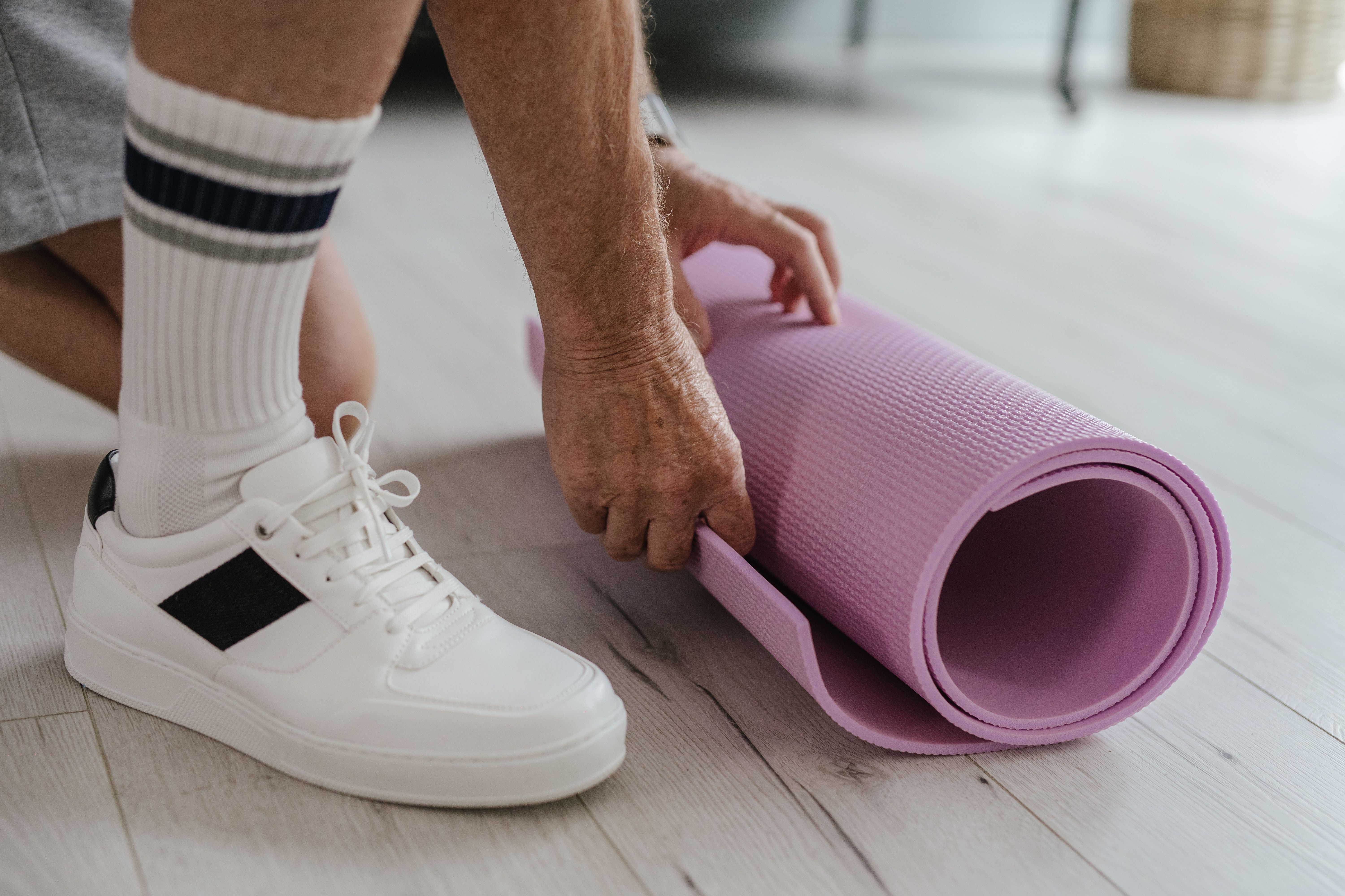 Close-up of a person rolling a pink yoga mat on a light wooden floor, wearing white sneakers.