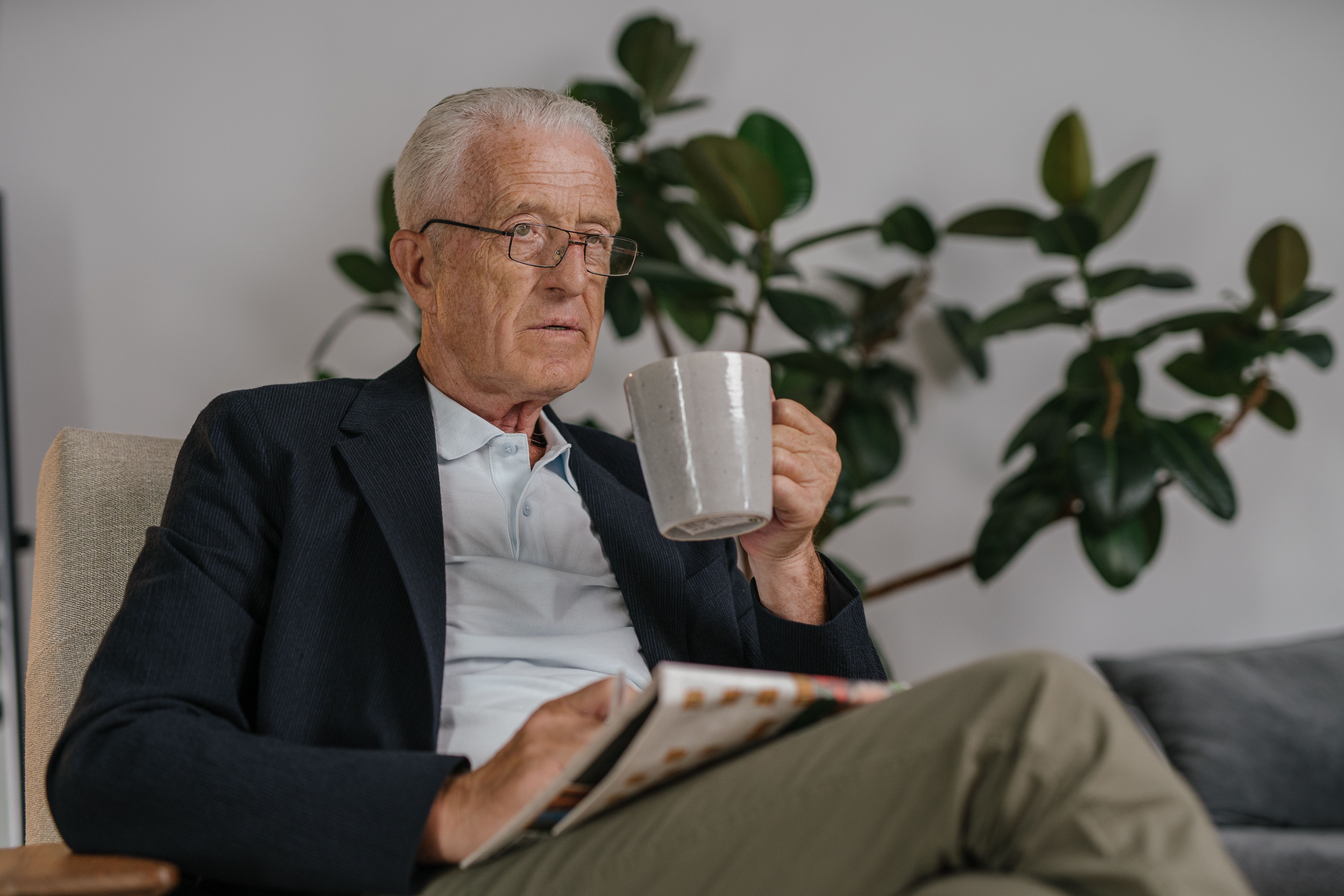 Elderly man in a black suit relaxing with a coffee mug indoors, looking relaxed and thoughtful.