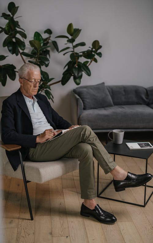 An Elderly Man in Black Suit Sitting on the Chair with His Legs Crossed while Reading Newspaper