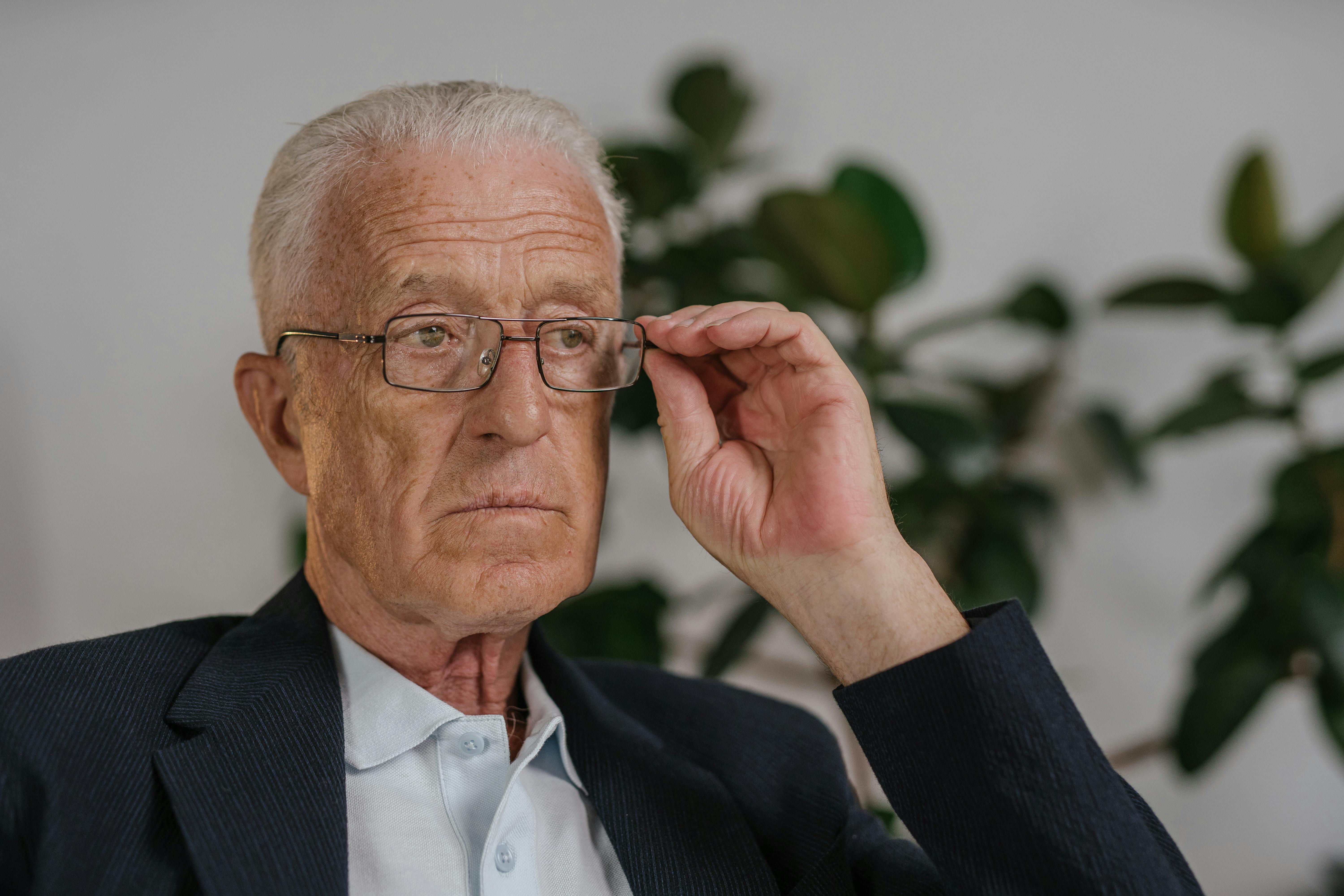Elderly man seated indoors, adjusting eyeglasses with a thoughtful expression.