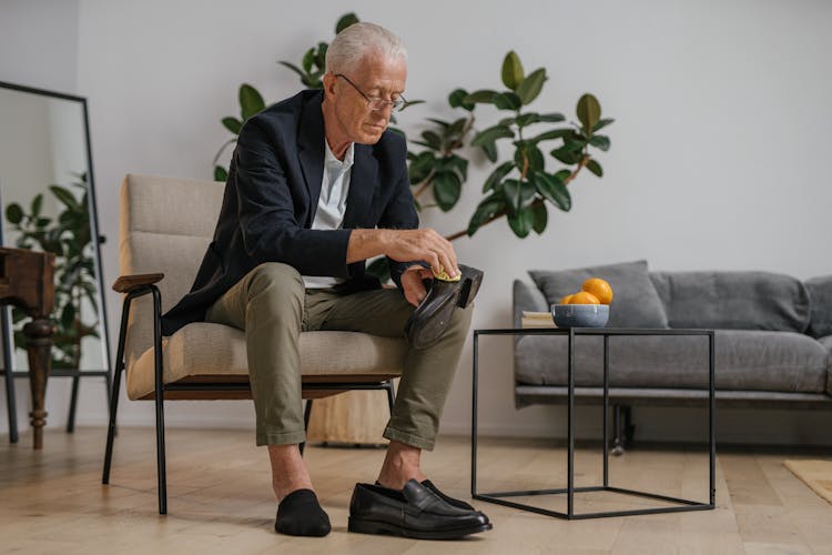 An Elderly Man In Black Suit Sitting On The Chair While Cleaning His Shoe