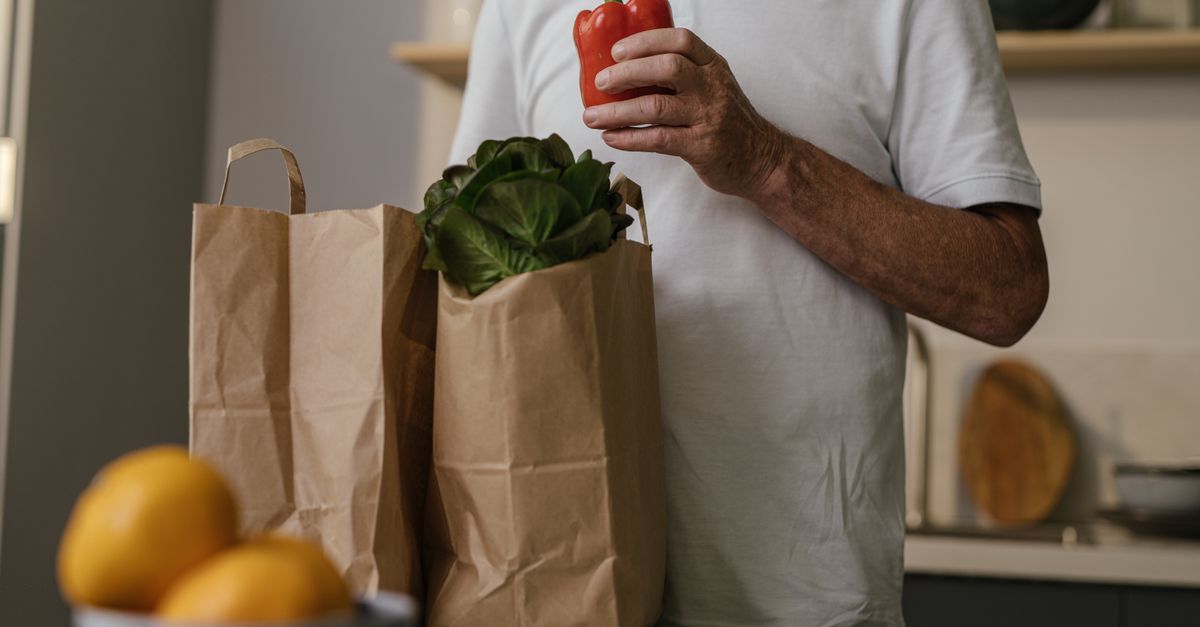 Homem idoso com compras, segurando um pimentão vermelho dentro de casa.