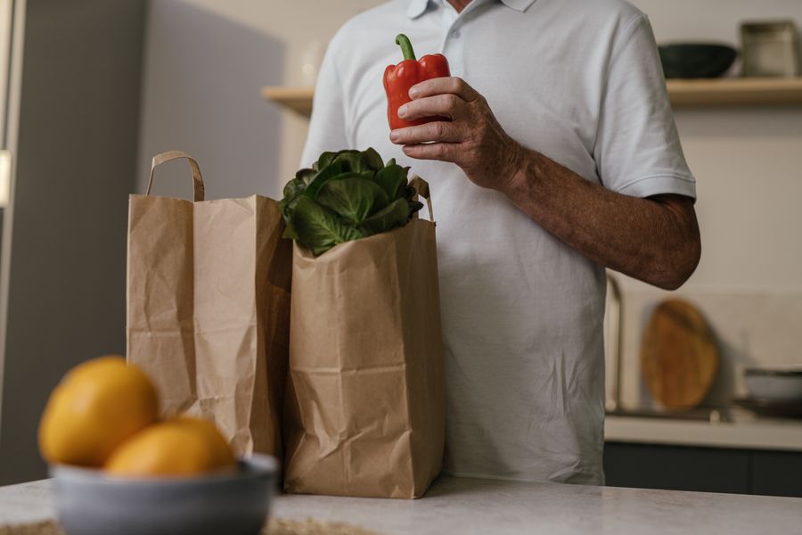 Elderly man with groceries, holding a red bell pepper indoors.