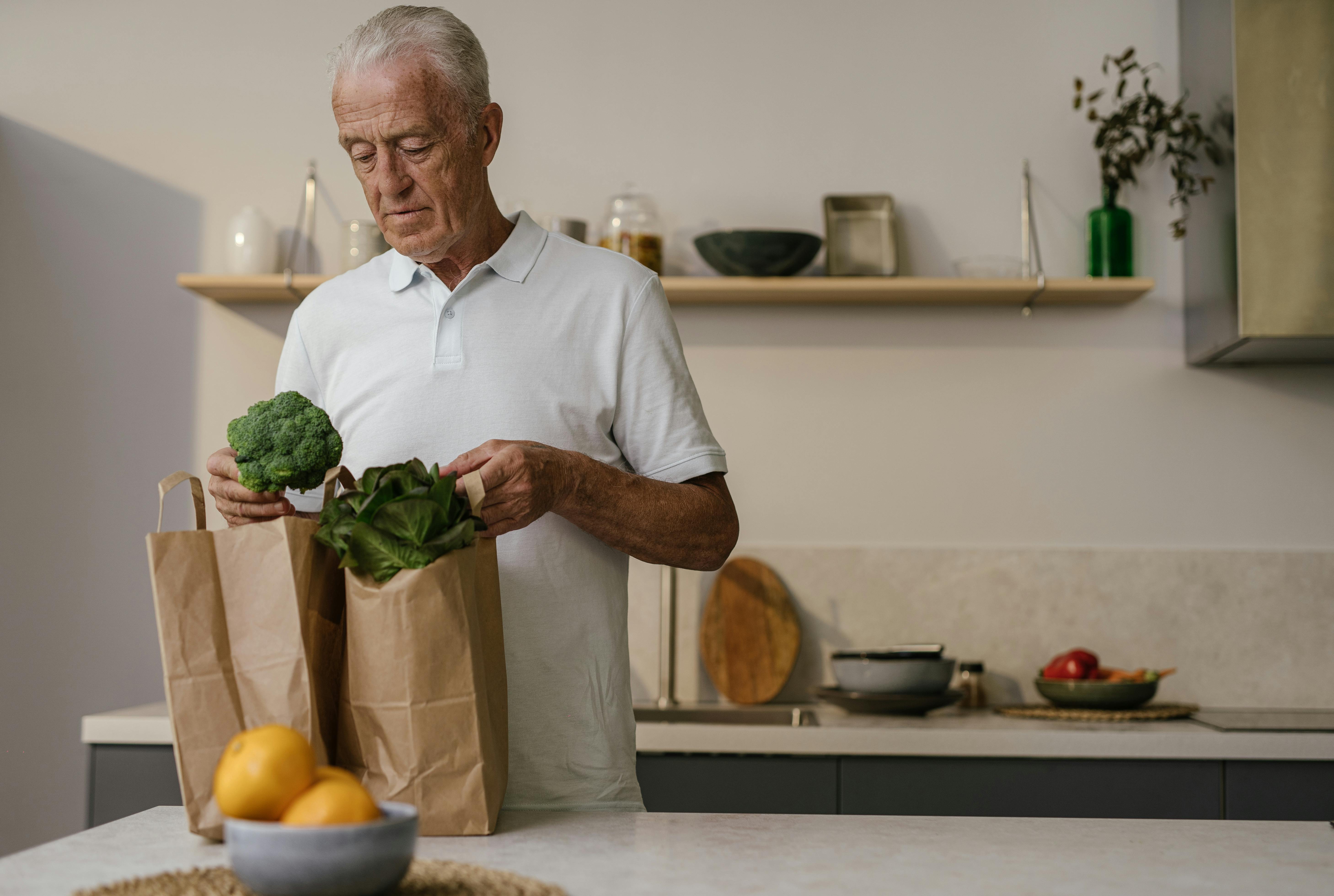 Elderly man in kitchen examining fresh vegetables from paper bags. Healthy lifestyle concept.