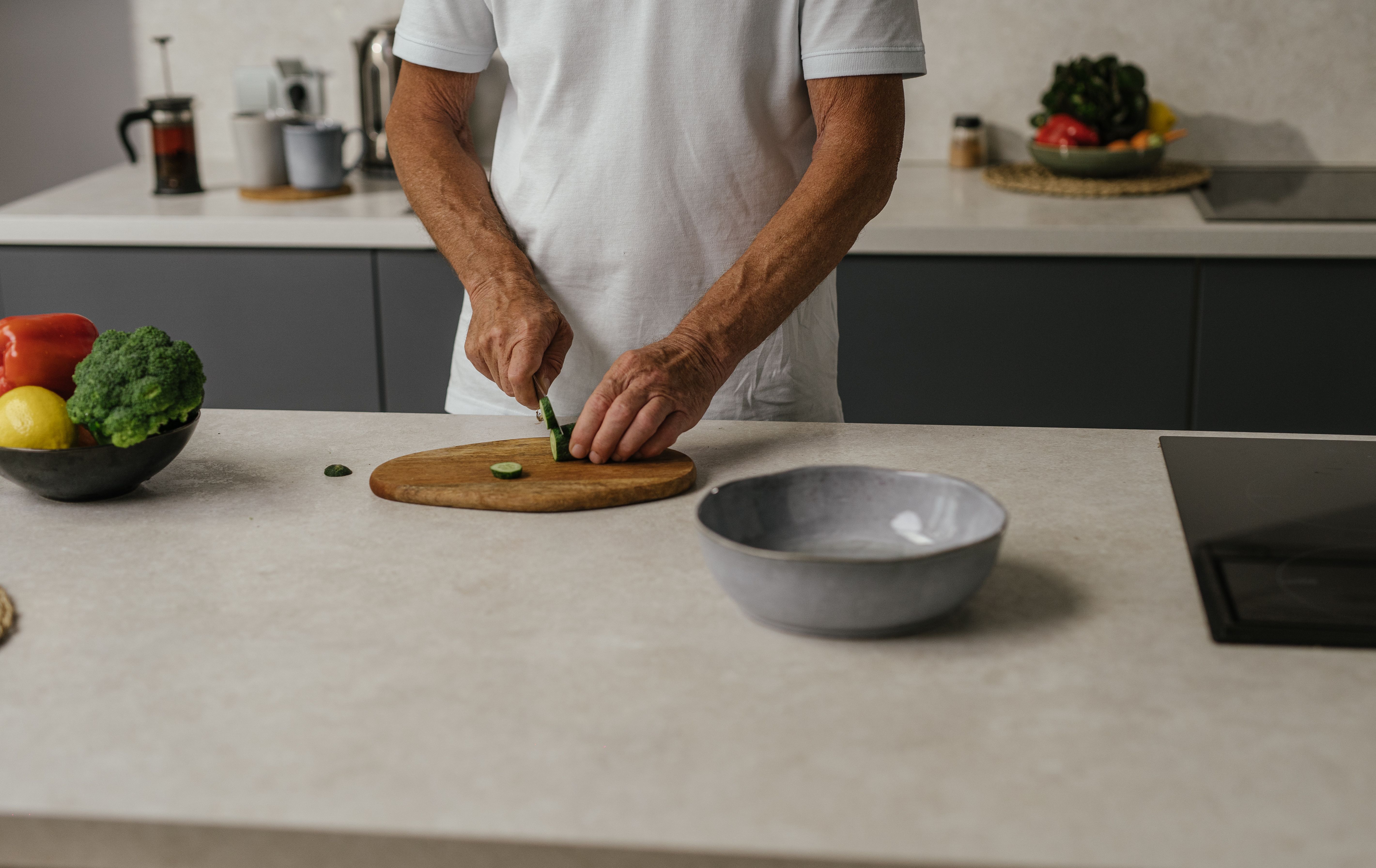 Elderly man slicing cucumber on a wooden board in a contemporary kitchen setting.