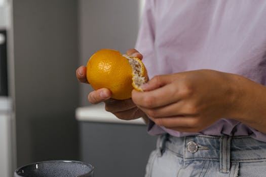 Close-up shot of hands peeling an orange indoors, highlighting fresh fruit texture and casual lifestyle.
