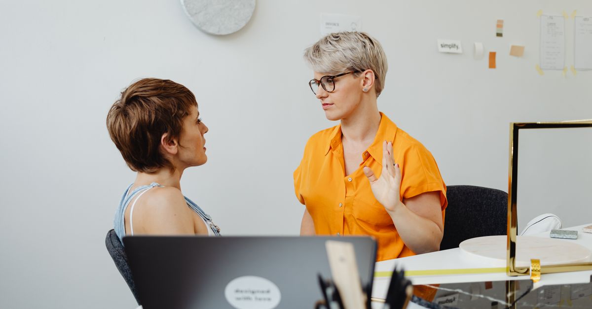 Two women having a discussion at a desk in a modern office setting, with laptops and office decor in the background.