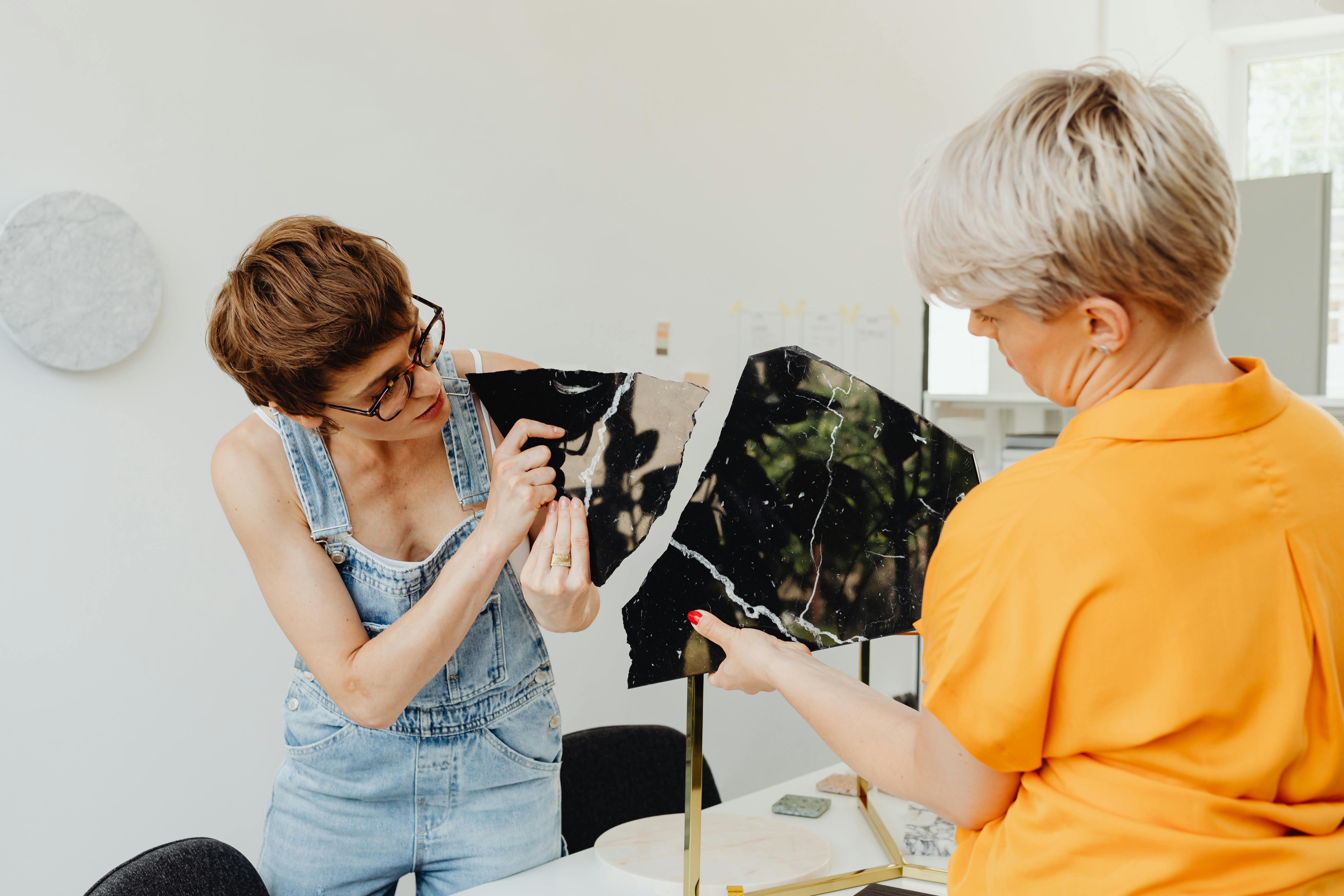 Two women examining broken marble tiles indoors, discussing repair strategies.