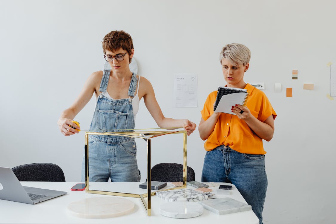 Woman in Blue Denim Overall Measuring Golden Table Stand Beside Woman in Yellow Shirt with Notebook