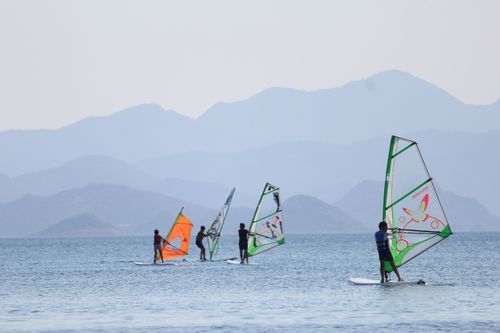 Free Four windsurfers ride on a tranquil sea with mountains in the background on a bright day. Stock Photo