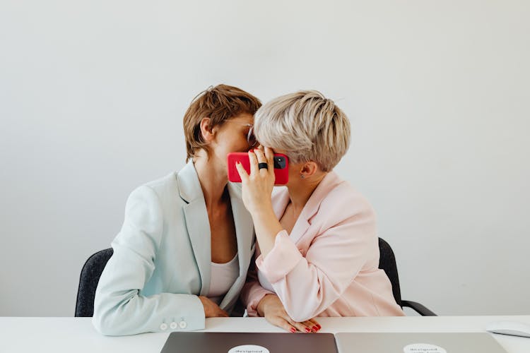 Women In Blazers Sitting Beside And Hiding A Kiss Behind Red Mobile Phone