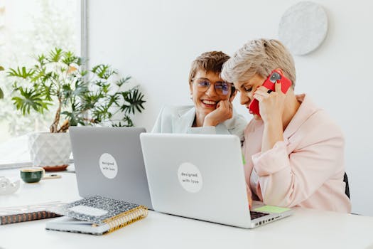 Two businesswomen working together in a bright office setting, using laptops and a mobile phone.