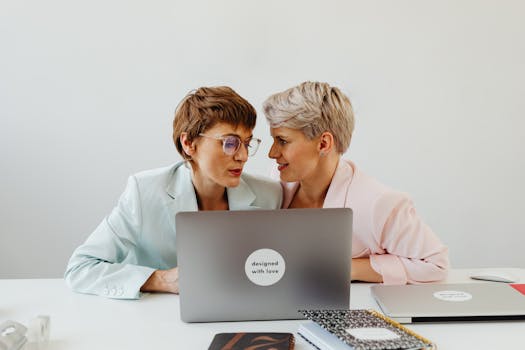 Two women in an office setting sharing a loving moment over work.