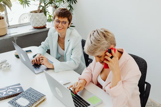Two women working together in a bright, modern office space, both using laptops and a phone.